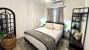 Ein Schlafzimmer mit Bett, Deckenventilator, Klimaanlage, einem Fenster mit Vorhängen, einem Nachttisch mit Lampe und einem Regal mit Pflanze.