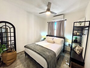 Un dormitorio con cama, ventilador de techo, unidad de aire acondicionado, ventana con cortinas, mesita de noche con lámpara y estante con una planta.