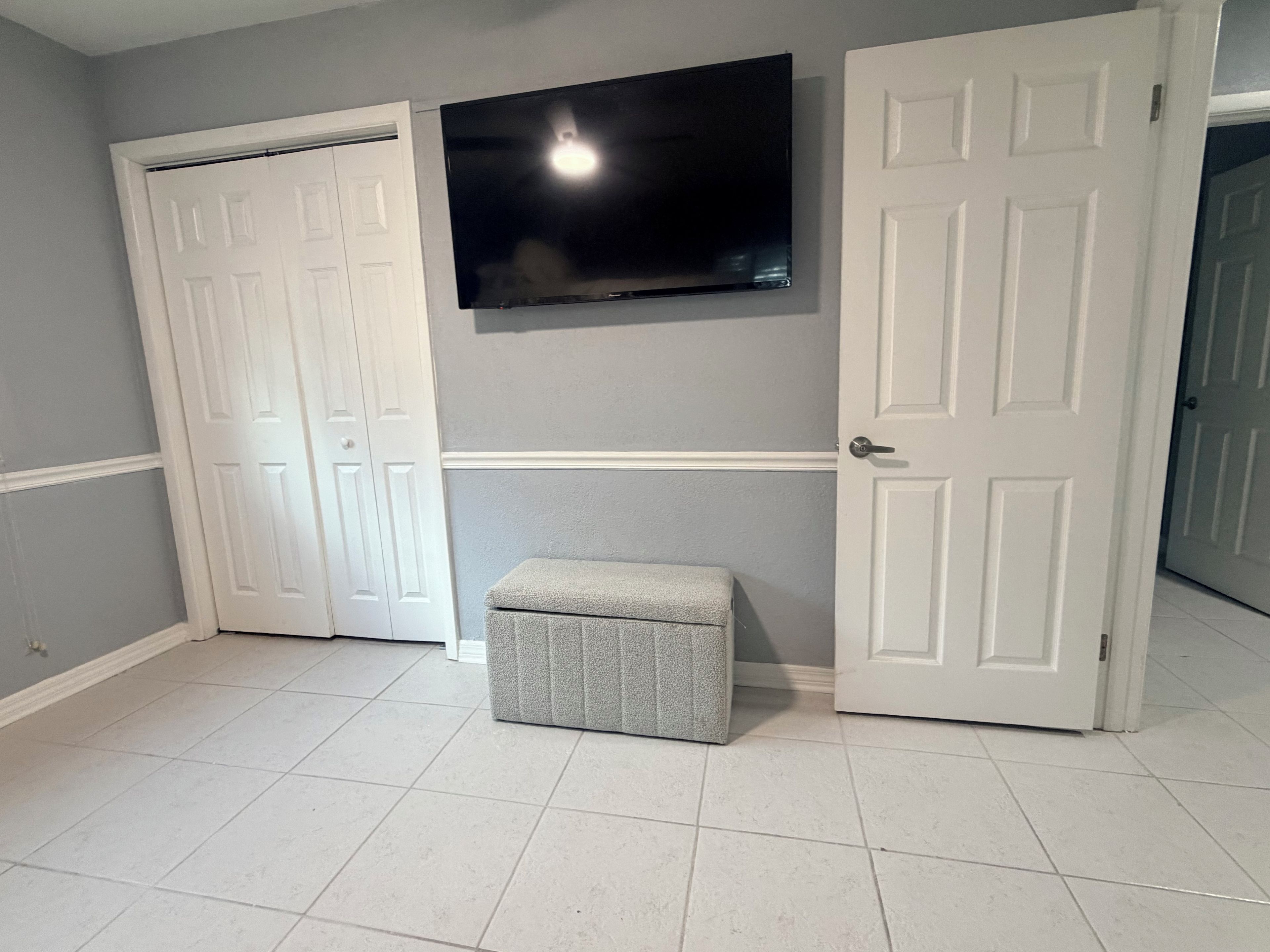 Un pasillo con una televisión de pantalla plana montada en la pared, una puerta blanca cerrada y un banco gris.