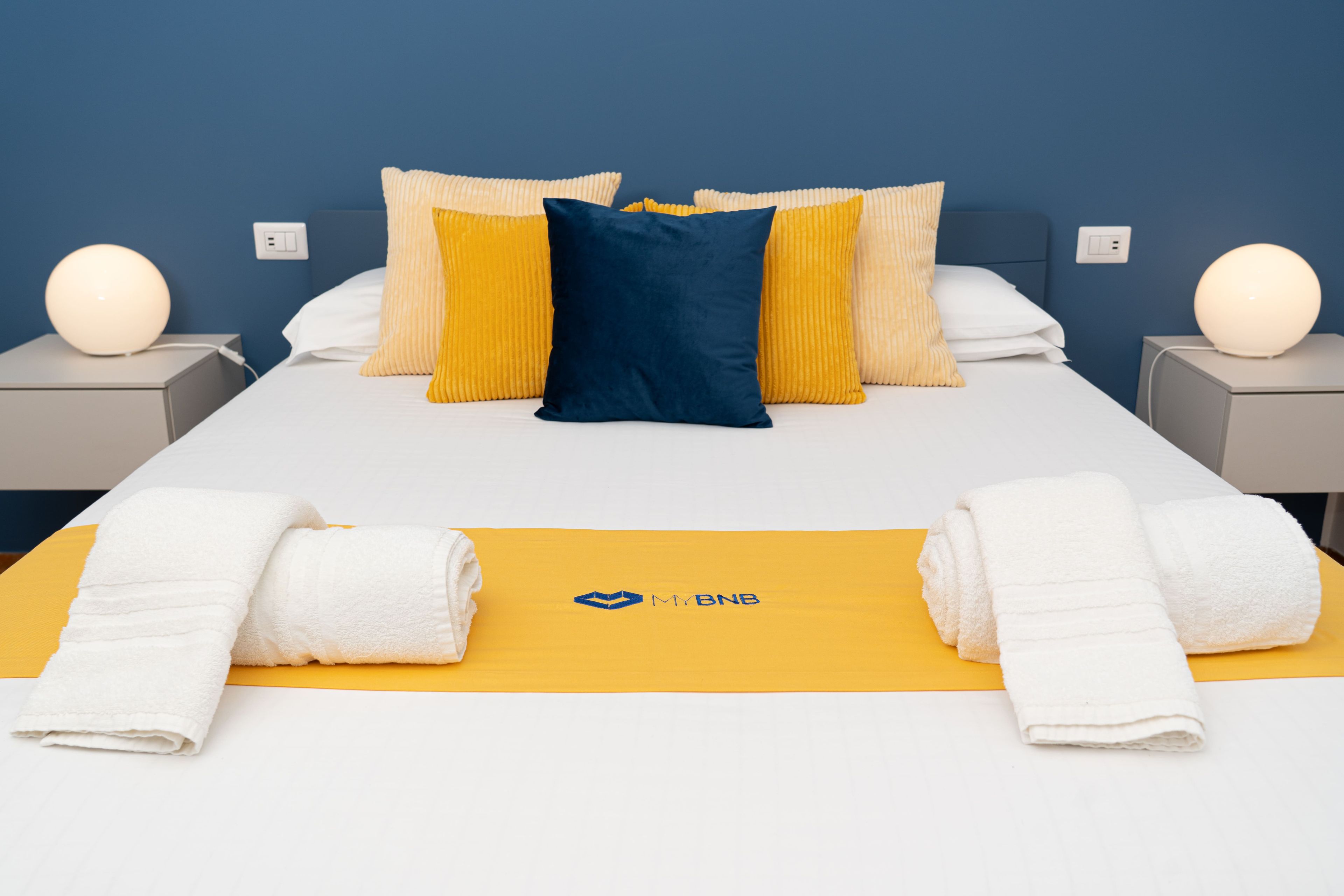 A neatly made bed with white and yellow bedding, a blue pillow, and a logo on the bedspread.