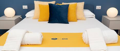 A neatly made bed with white and yellow bedding, a blue pillow, and a logo on the bedspread.