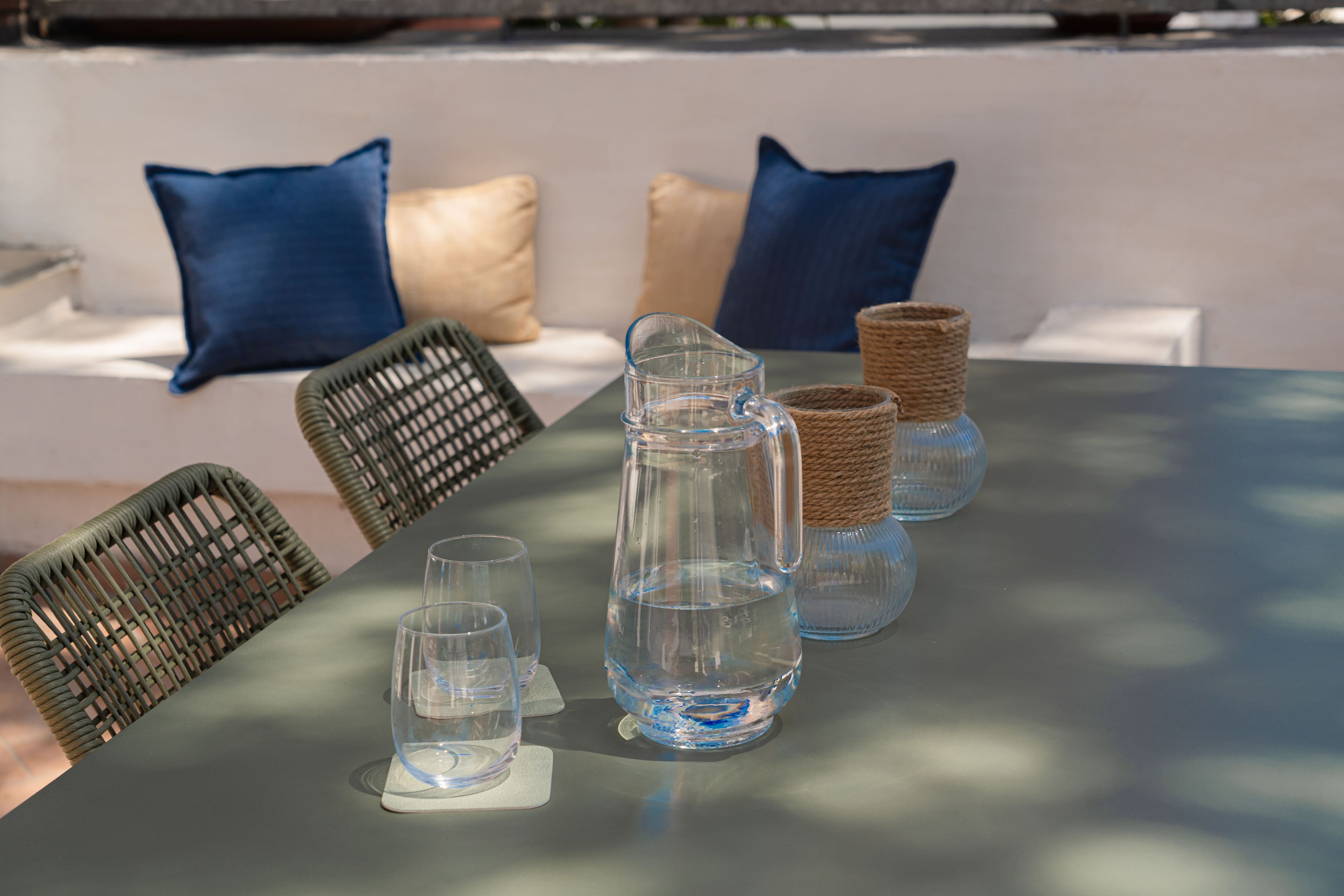 A table with clear glassware and a pitcher, set against a white wall with blue cushions.