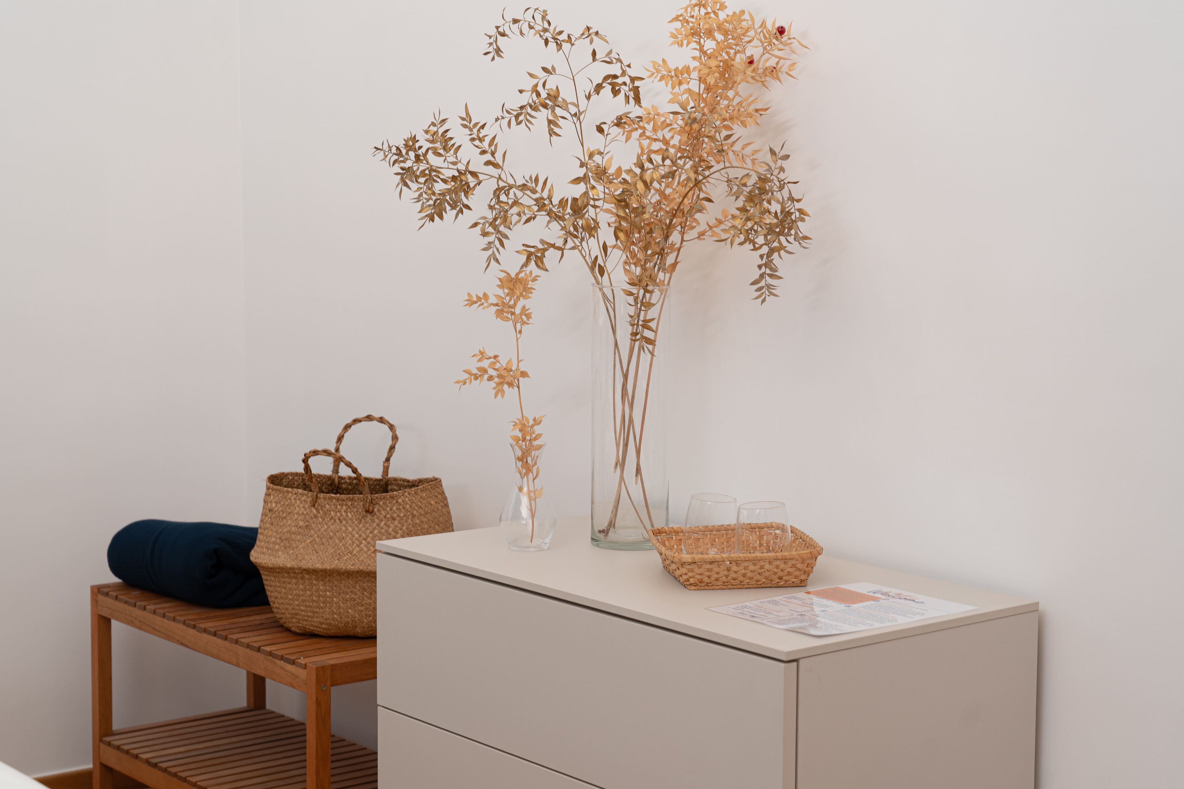 Quarto com um banco de madeira, um armário branco e uma prateleira com galhos decorativos e uma cesta trançada.