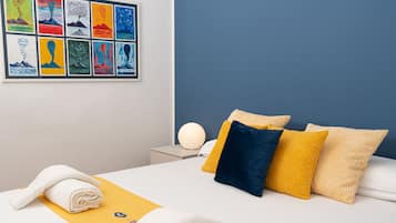 Quarto de hotel com cama, almofadas, mesa de cabeceira com abajur e uma parede com obras de arte.