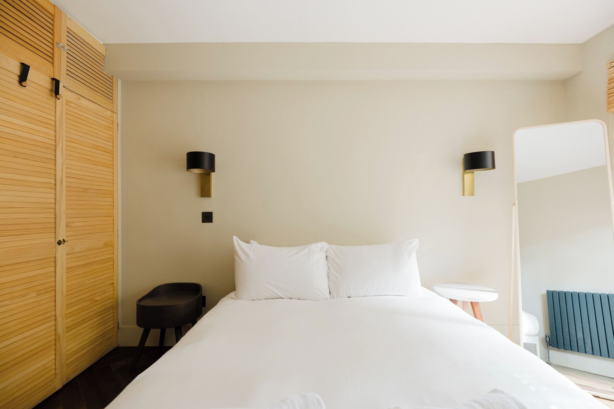 A hotel room with a bed, a small side table, a chair, a mirror, and a radiator.