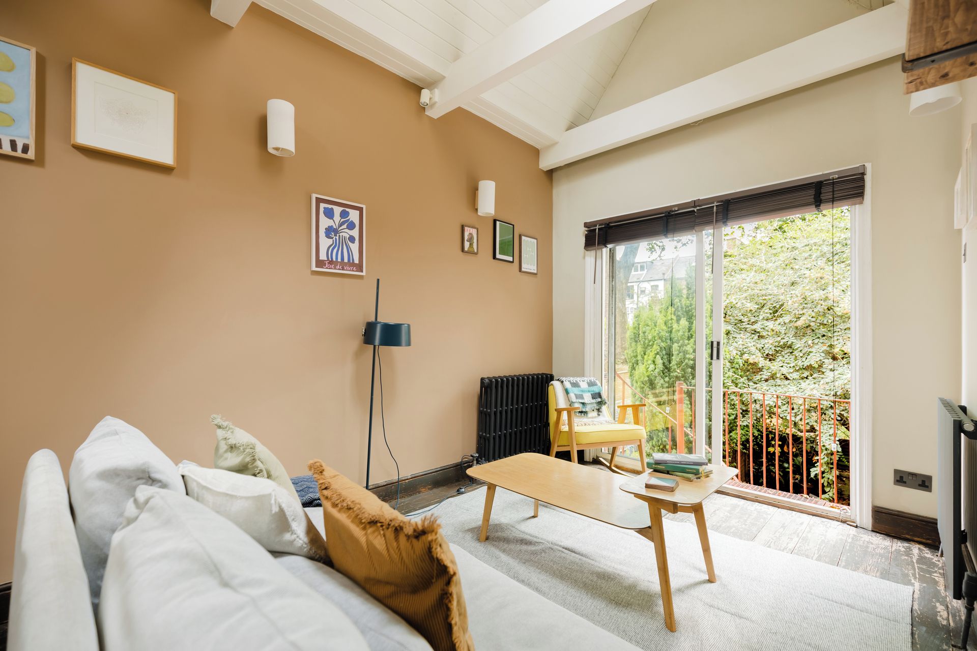 A living room with a sofa, a table, and a chair. There are framed pictures on the wall and a radiator.