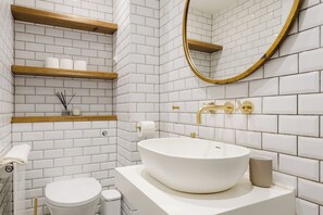 A modern bathroom with white subway tiles, a white sink with a gold faucet, and a wooden shelf holding toiletries. - Sublime 1BDR Coach House Conversion in Hackney (London)