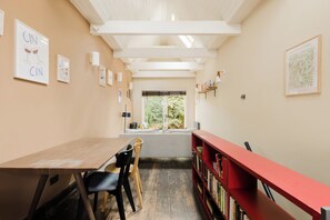 A room with a wooden table, chairs, a bookshelf, and a window with a view of greenery. - Sublime 1BDR Coach House Conversion in Hackney (London)