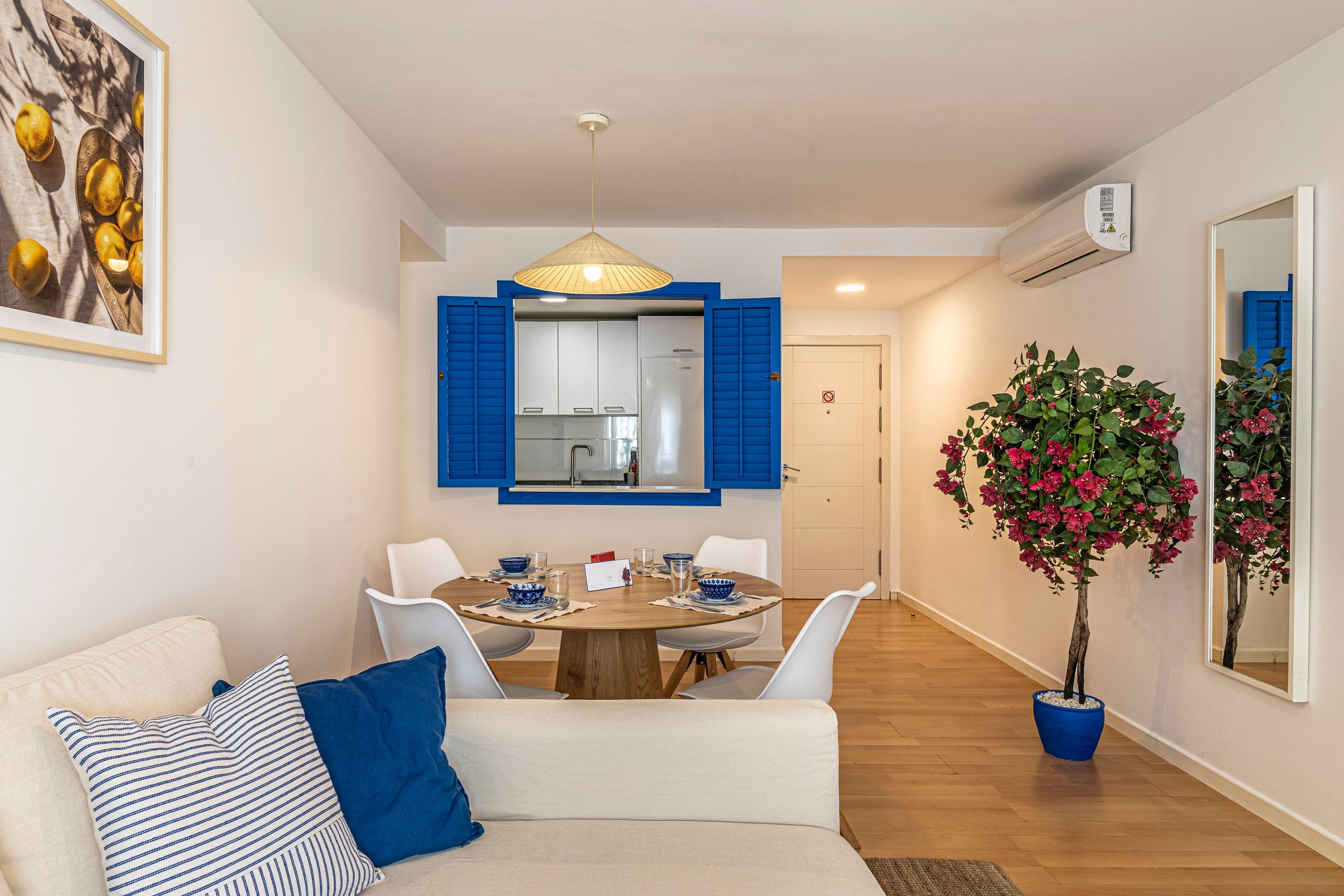Een moderne woonkamer met een eethoek, een bank met blauwe kussens, een potplant en een zichtbare keuken door een open deur.