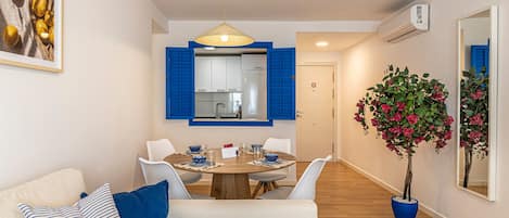 A modern living room with a dining area, a sofa with blue cushions, a potted plant, and a kitchen visible through an open door.