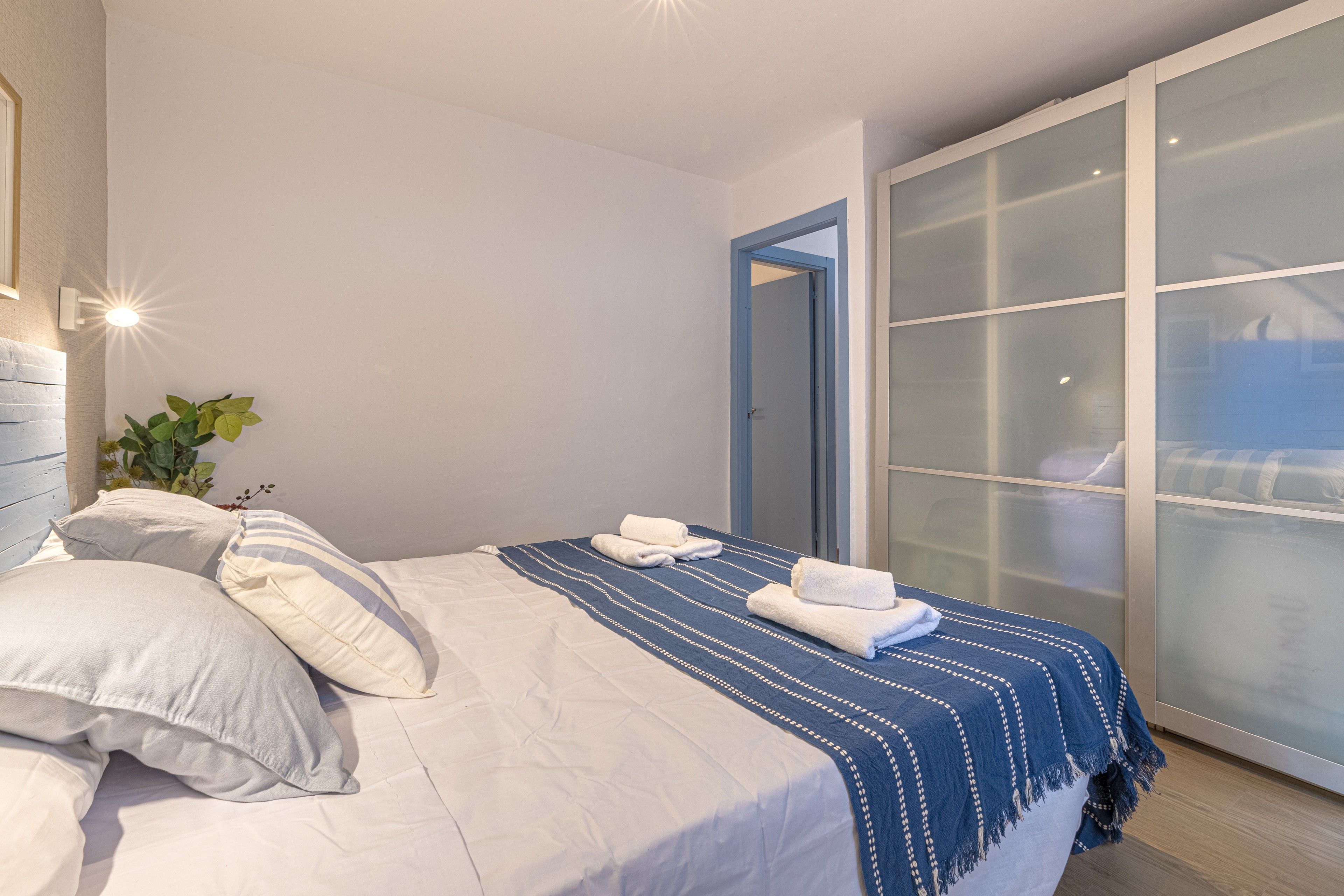 Una cama bien hecha con almohadas y una manta azul a rayas, una planta en maceta y un ventanal corredera que da a otra habitación.