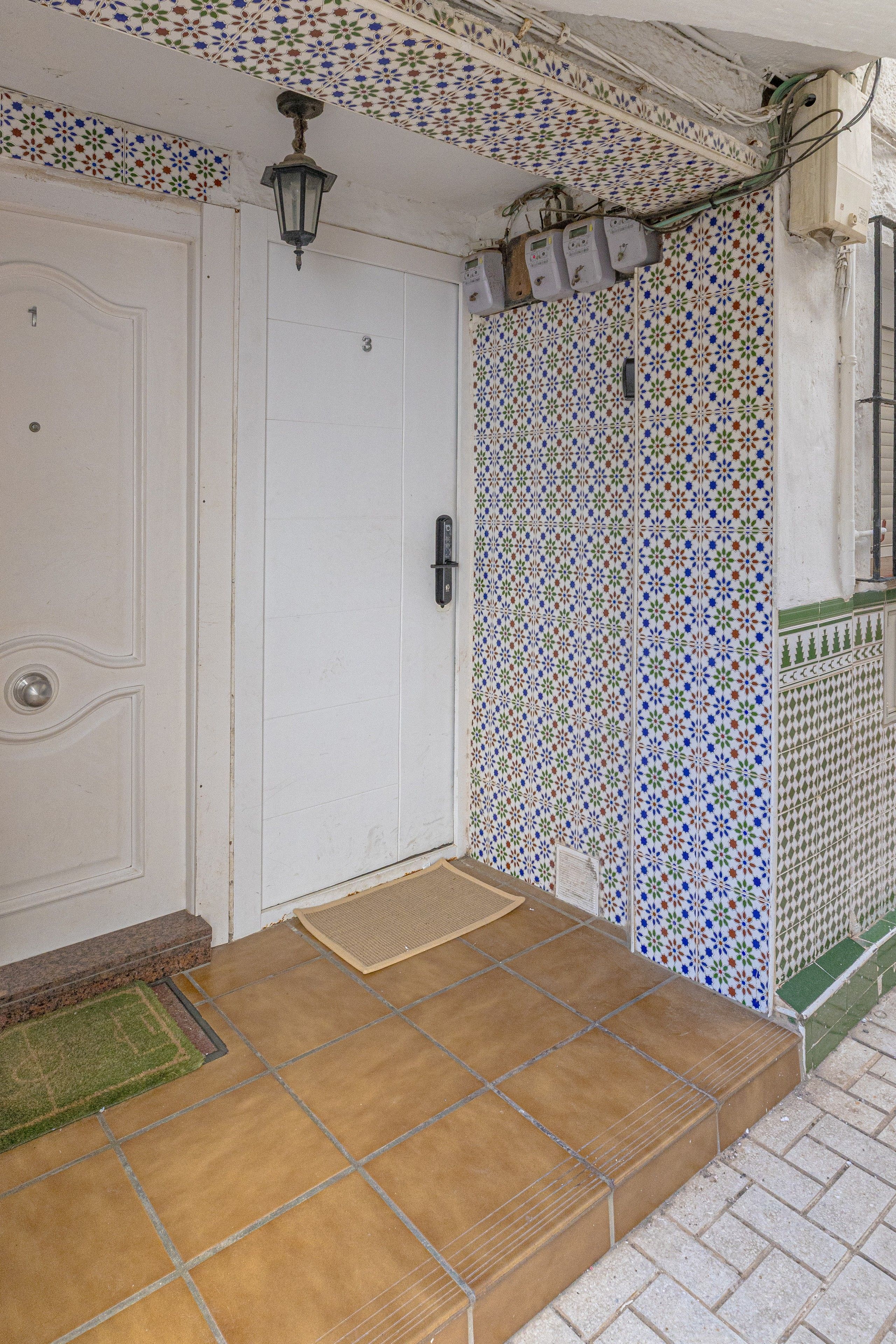 Una puerta blanca con una placa de número, un vestíbulo con baldosas y un felpudo, y una pared con baldosas y un diseño decorativo.