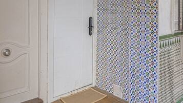 Una puerta blanca con una placa de número, un vestíbulo con baldosas y un felpudo, y una pared con baldosas y un diseño decorativo.