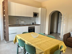 Een kleine keuken met witte kastjes, een magnetron en een spoelbak. Er staat een eettafel met een gele tafelkleed en groene stoelen.