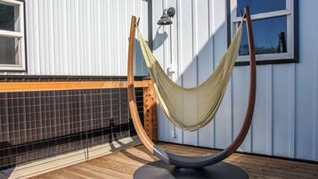 Un fauteuil suspendu, de type hamac, sur une terrasse en bois, avec une clôture en métal et un bâtiment en arrière-plan.