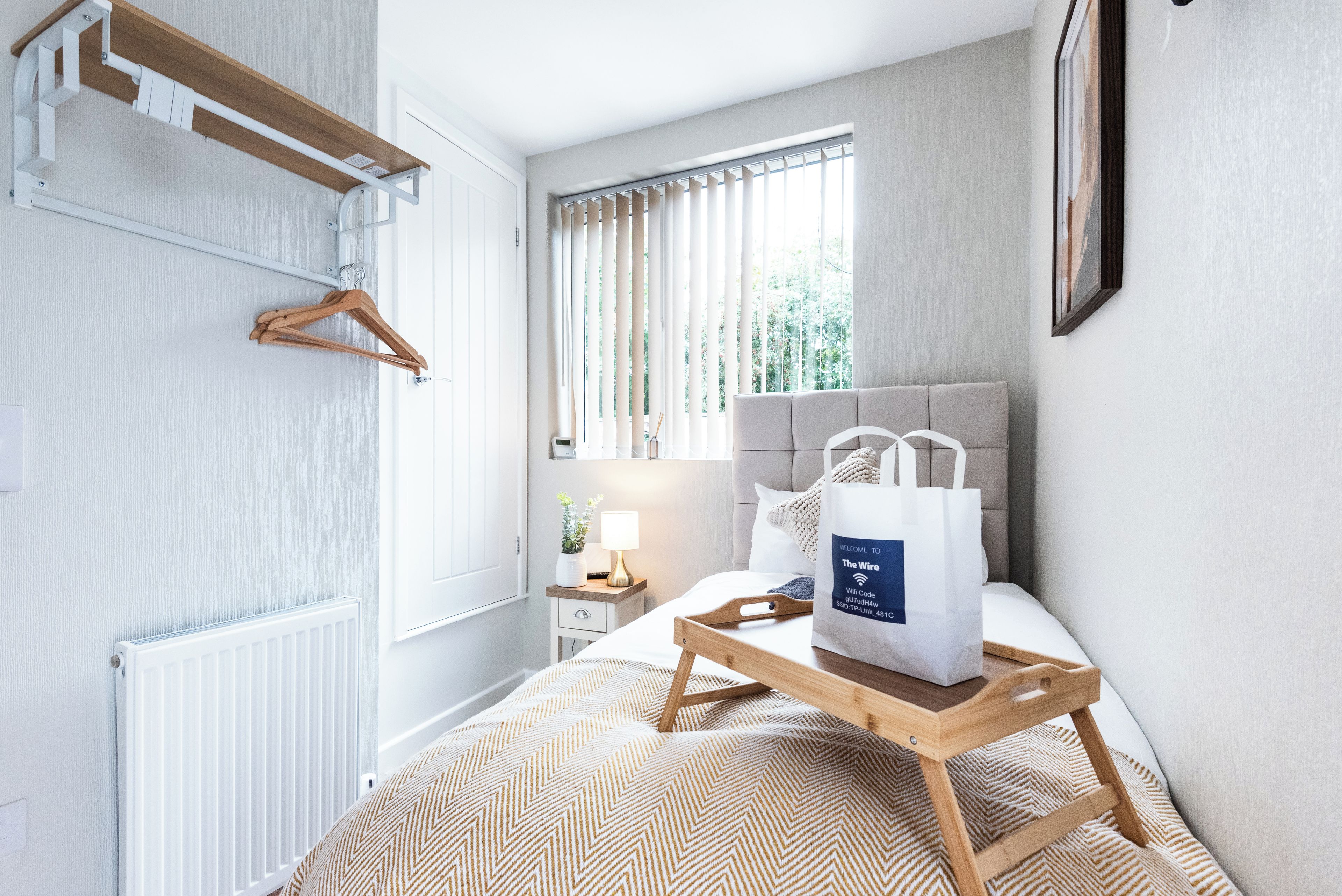 Habitación de hotel con cama, mesita de noche con lámpara, perchero y ventana con cortinas.