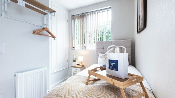 Habitación de hotel con cama, mesita de noche con lámpara, perchero y ventana con cortinas.