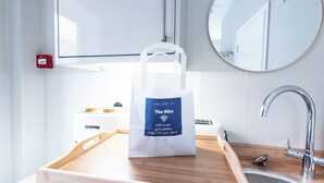 Habitación de hotel con una bandeja de madera que contiene una bolsa de bienvenida, un lavamanos, un espejo y un dispositivo GEEPAS.