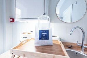 Une chambre d’hôtel avec un plateau en bois contenant un sac de bienvenue, un lavabo, un miroir et un appareil GEEPAS.