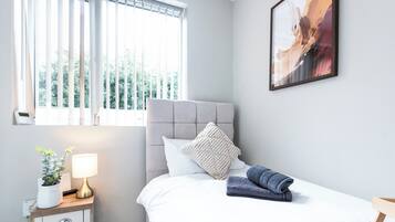 A bedroom with a bed, a bedside table, a lamp, a plant, and a window with blinds.