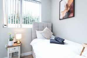 Ein Schlafzimmer mit einem Bett, einem Nachttisch, einer Lampe, einer Pflanze und einem Fenster mit Jalousien.
