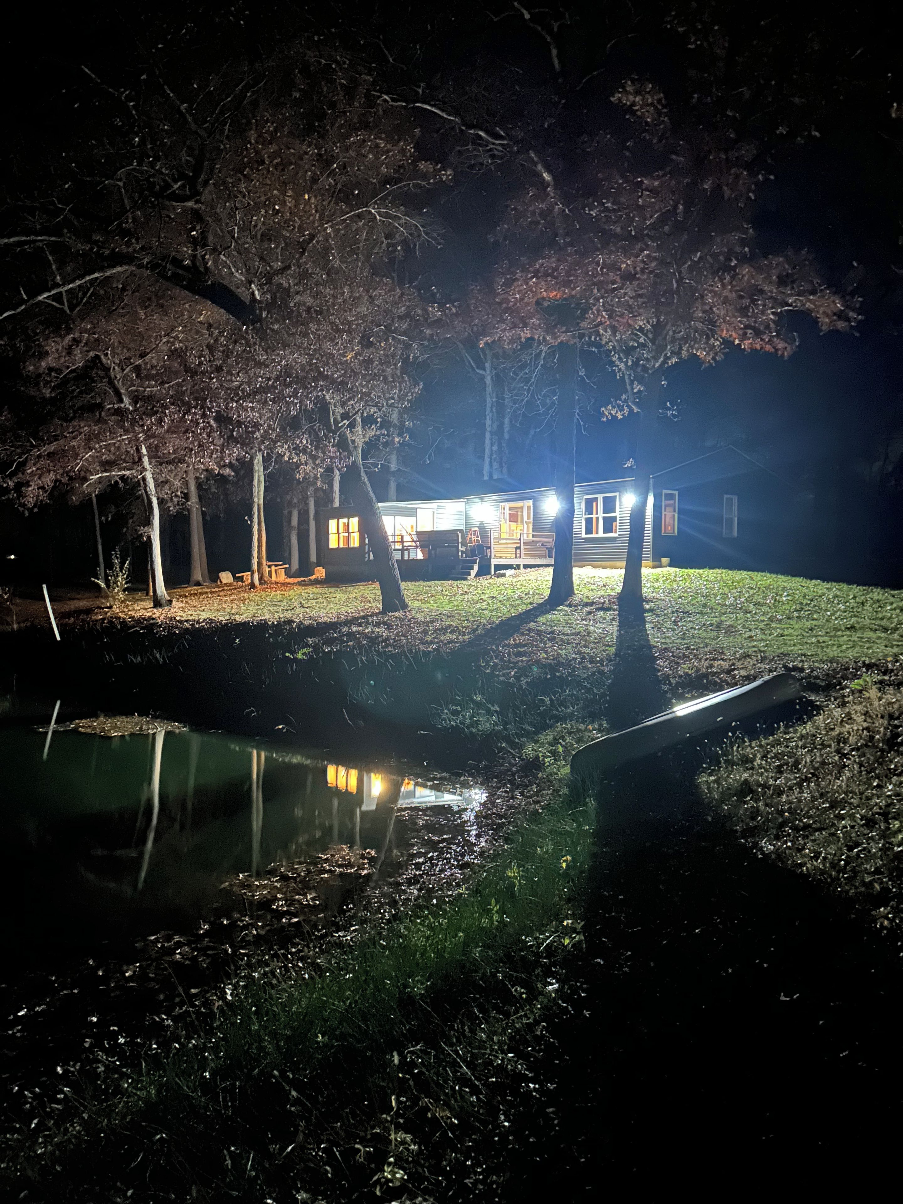 A house with a pond in front, surrounded by trees and a well-lit yard at night.