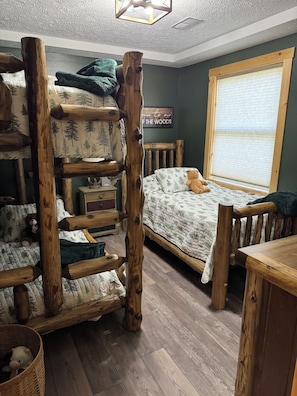 A room with a bunk bed, a wooden ladder, a window with blinds, a bedside table, and a wall sign.