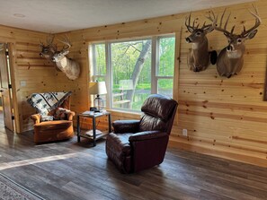 Interior de una cabaña con paredes de madera, un sillón de cuero, una mesa con lámpara y cornamentas de ciervo colgadas.