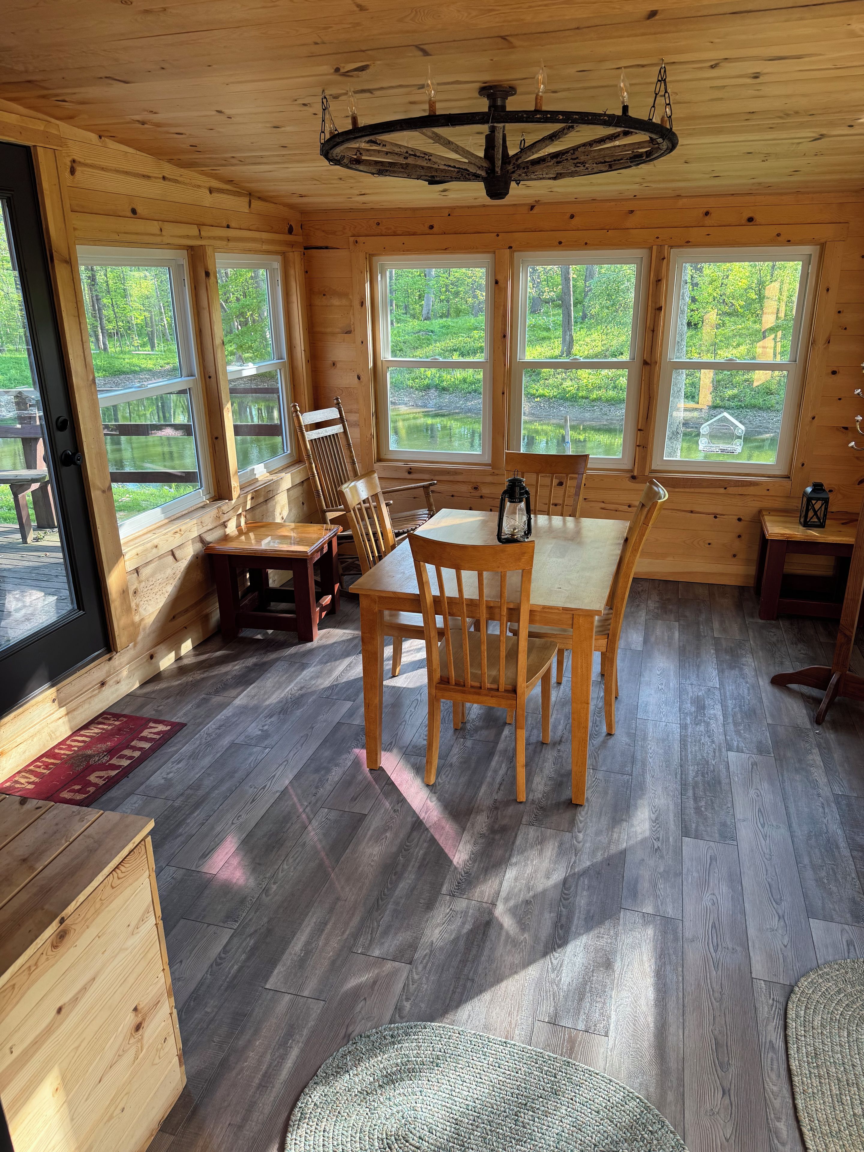 A wooden cabin interior with a dining table, chairs, a lantern, and large windows overlooking a natural landscape.
