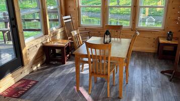 A wooden cabin interior with a dining table, chairs, a lantern, and large windows overlooking a natural landscape.