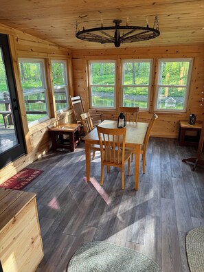 A wooden cabin interior with a dining table, chairs, a lantern, and large windows overlooking a natural landscape.