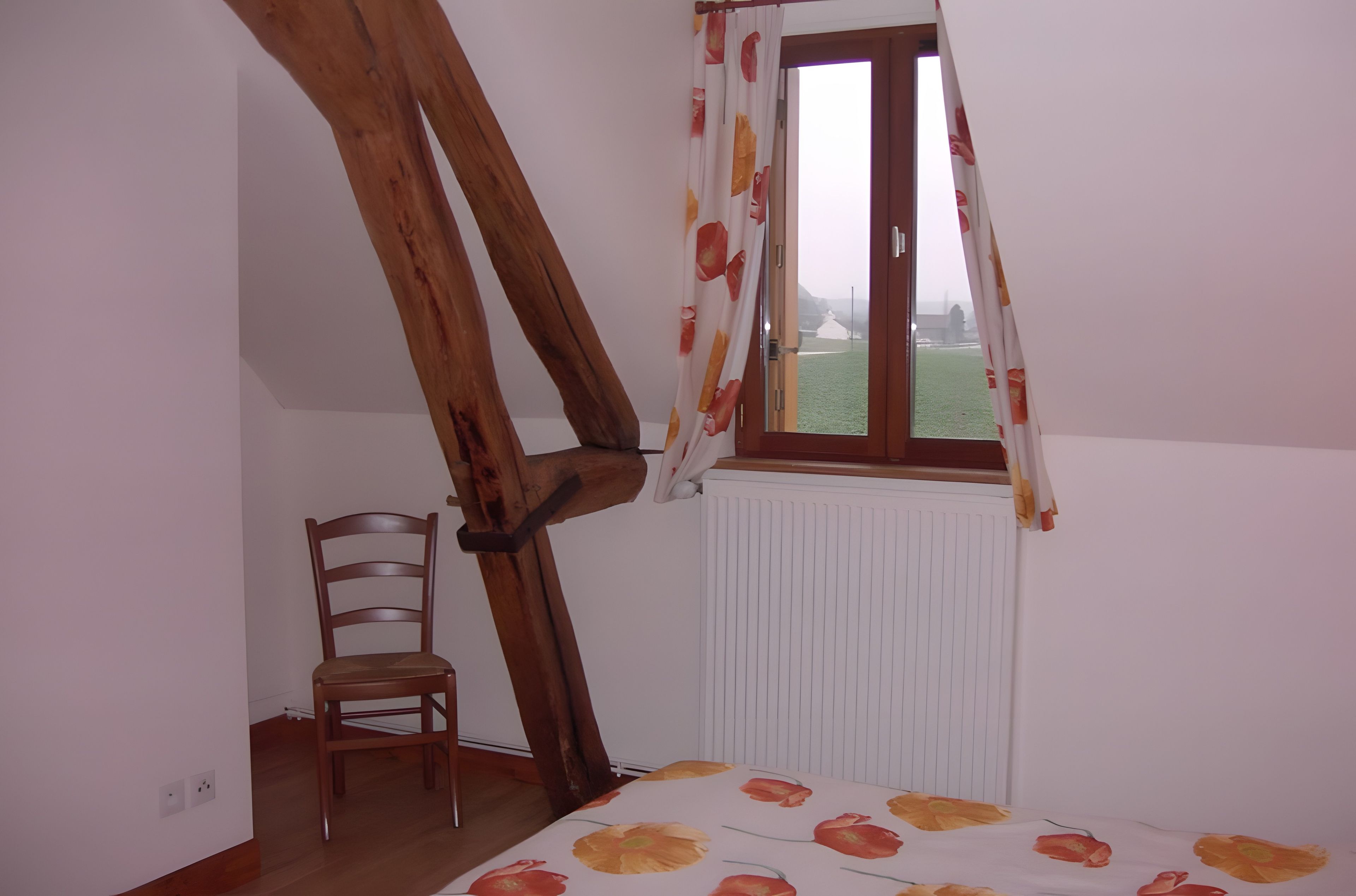 Une pièce avec un plafond en pente, une chaise en bois, un radiateur et une fenêtre avec des rideaux.