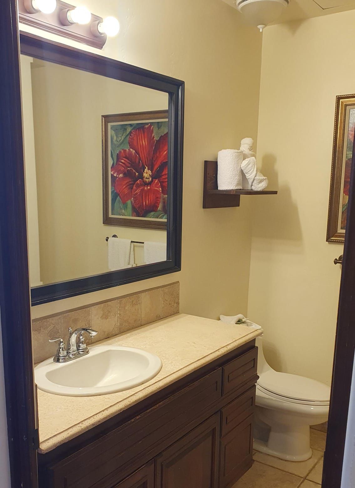 Een badkamer met een grote spiegel, een wastafel, een toilet en een lijst met een bloemistisch schilderij aan de muur.