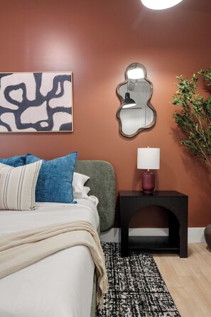 Ein Schlafzimmer mit Bett, Nachttisch mit Lampe, Spiegel und einem an der Wand hängenden abstrakten Gemälde.
