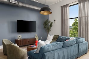 Een moderne woonkamer met een grote flatscreen televisie, een blauwe bank, een houten kast en een groot raam met gordijnen.