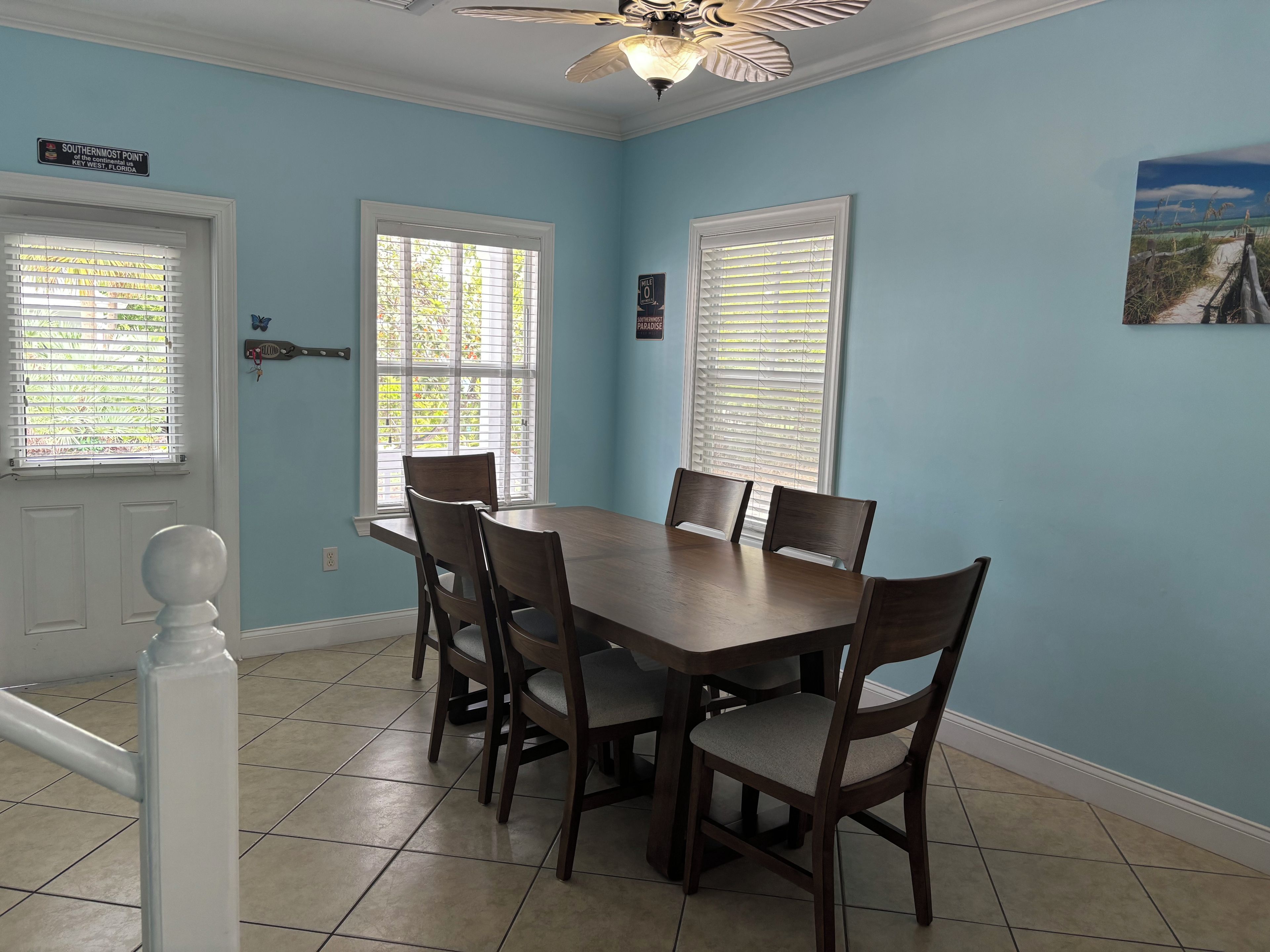 A dining room with a wooden table and chairs, two windows with blinds, a ceiling fan, and a door with a sign.