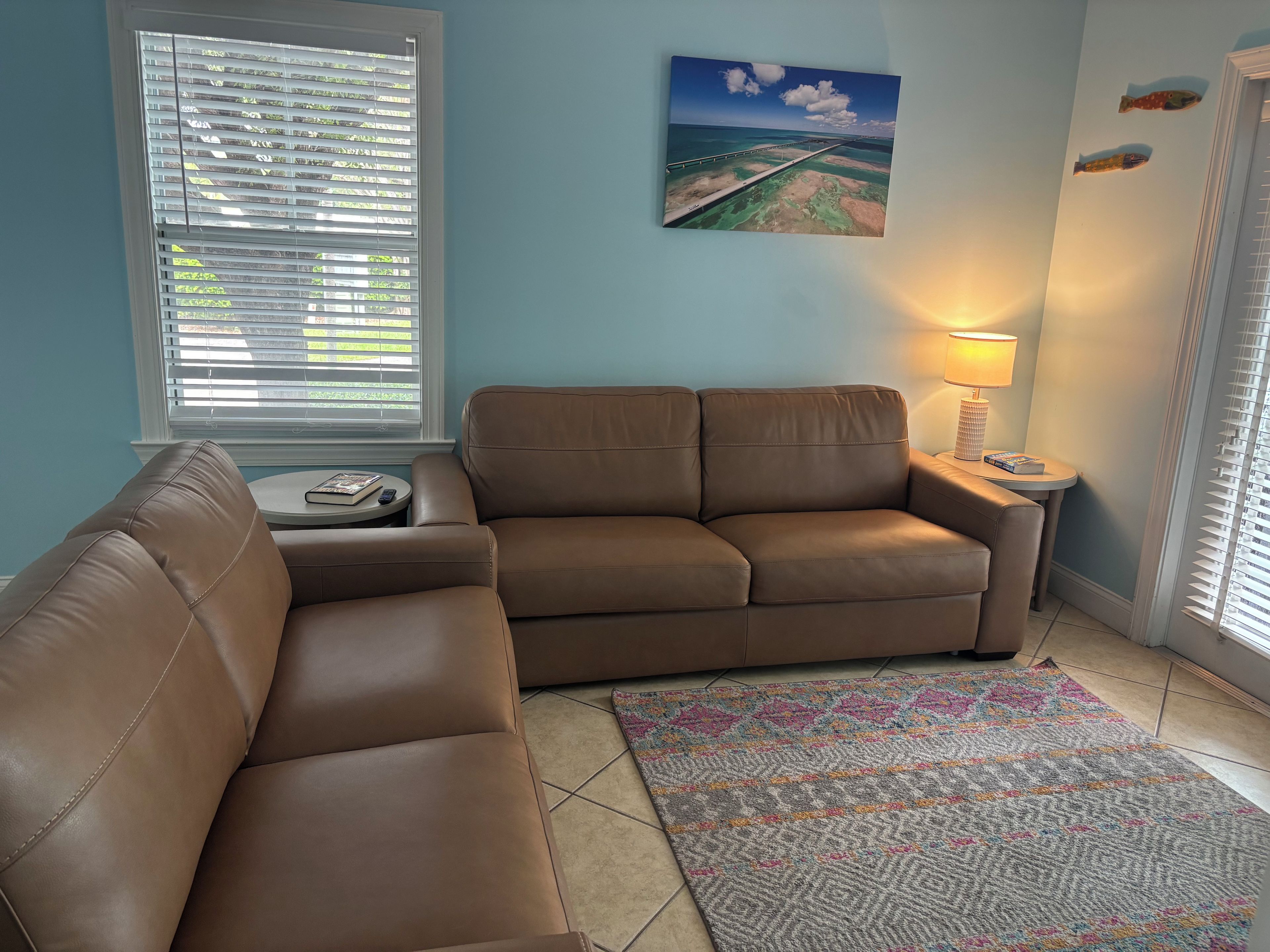 A living room with a brown leather sofa, a patterned rug, a painting of a bridge, and a lamp on a side table.