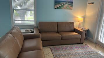 A living room with a brown leather sofa, a patterned rug, a painting of a bridge, and a lamp on a side table.