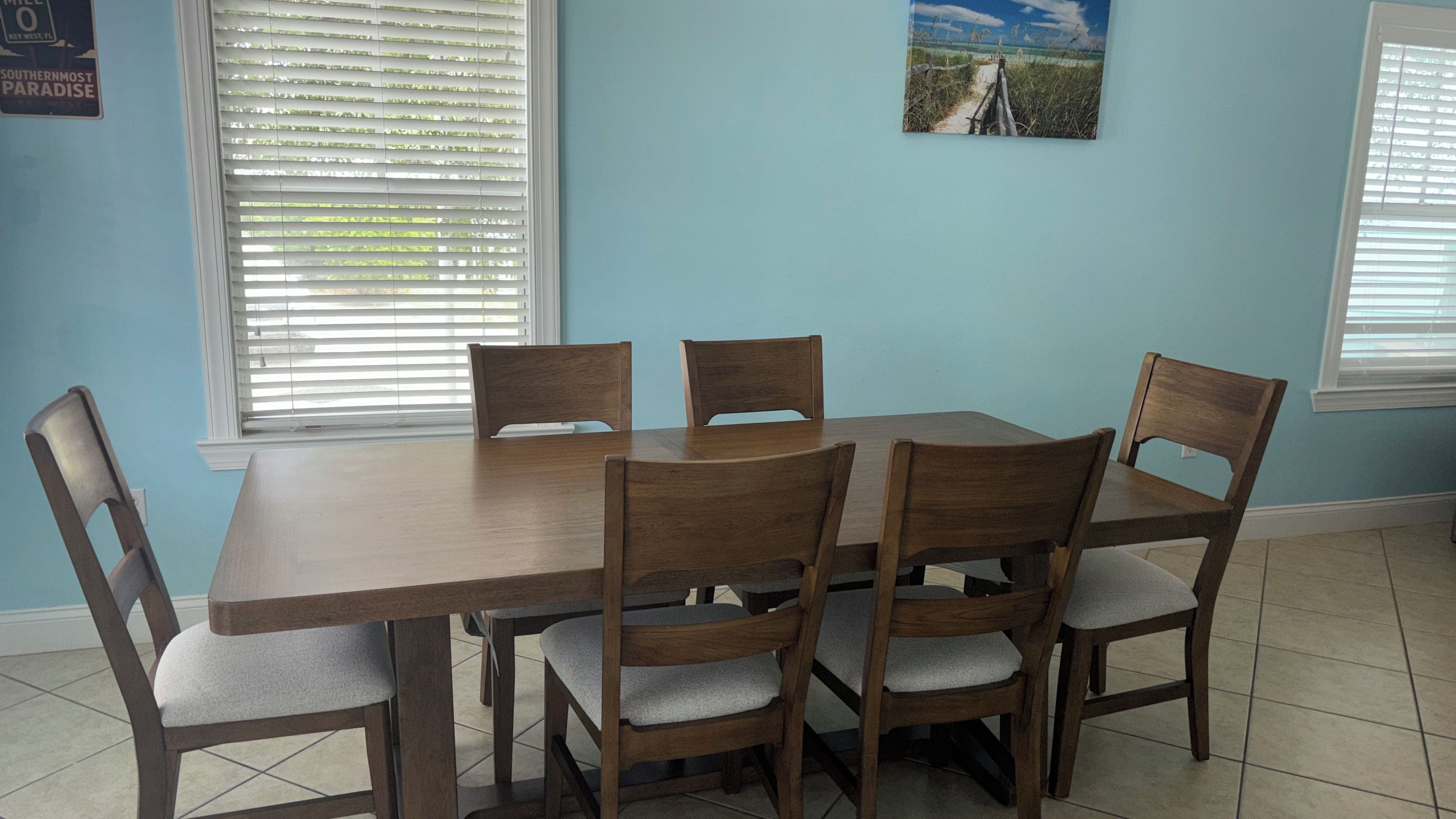 Un comedor con una mesa de madera y seis sillas, dos ventanas con persianas y un cuadro en la pared.