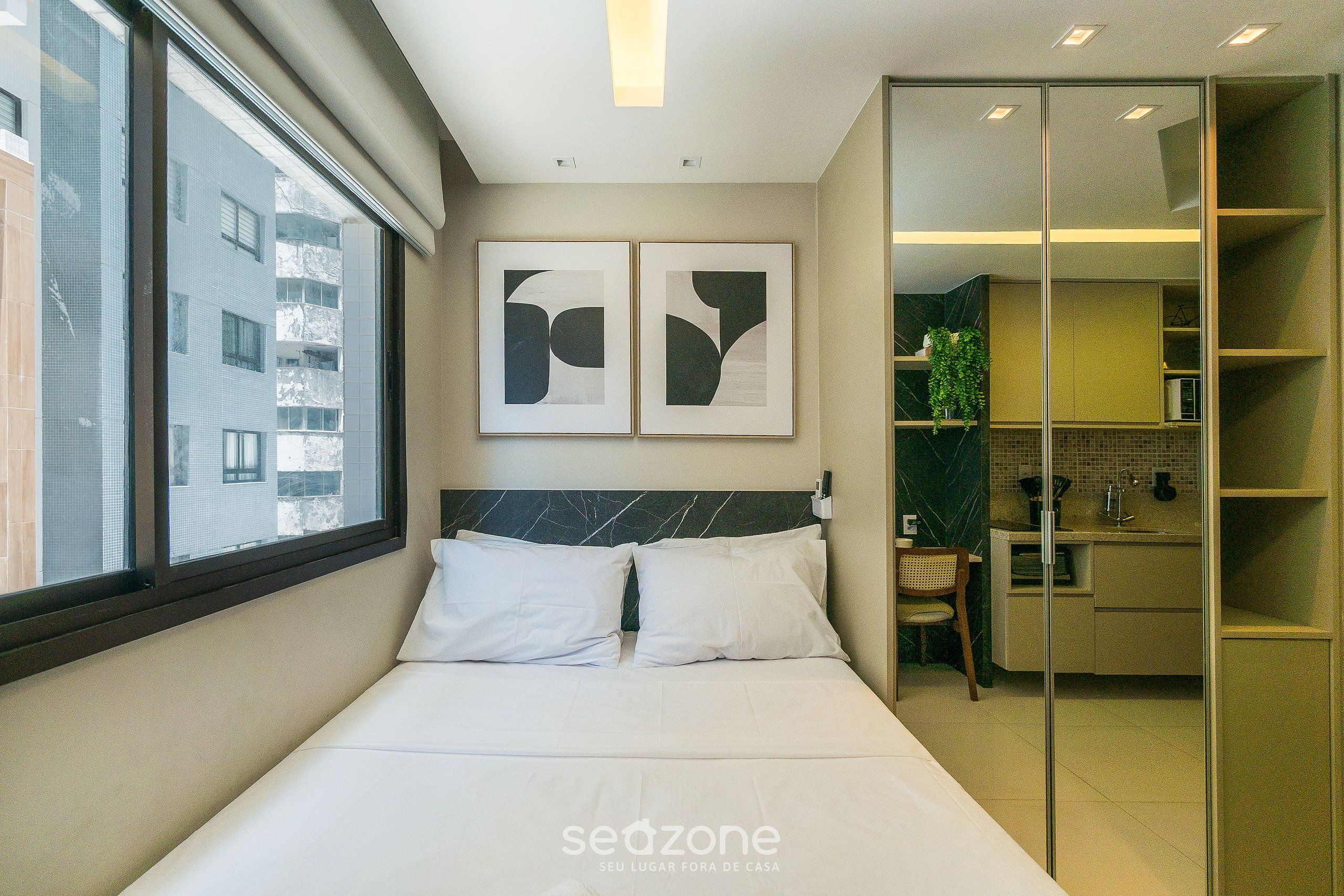 A modern hotel room with a bed, two wall-mounted artworks, a view of the outside, and a kitchenette area.