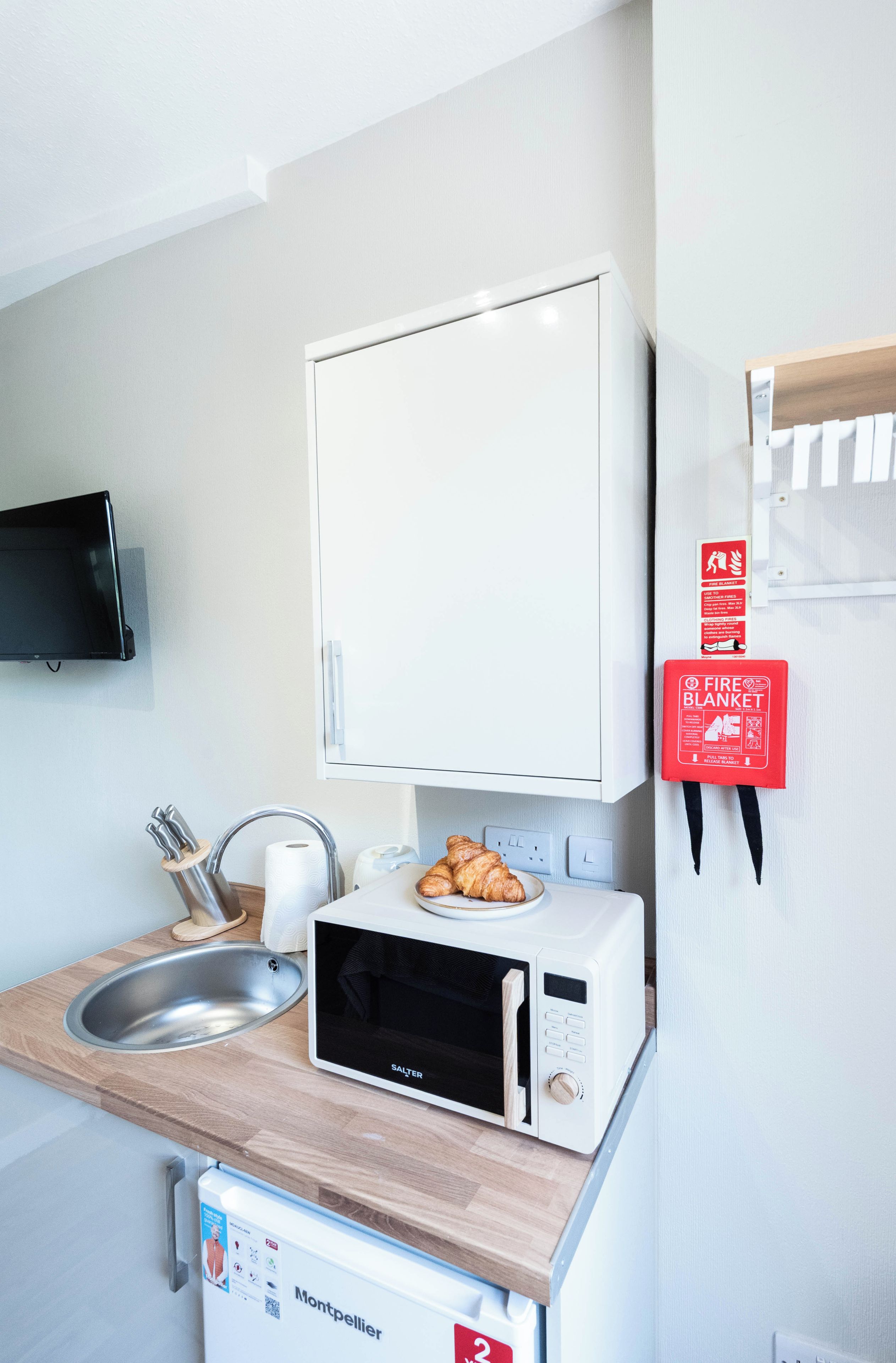 A compact kitchenette with a microwave, a small sink, and a fire blanket mounted on the wall.