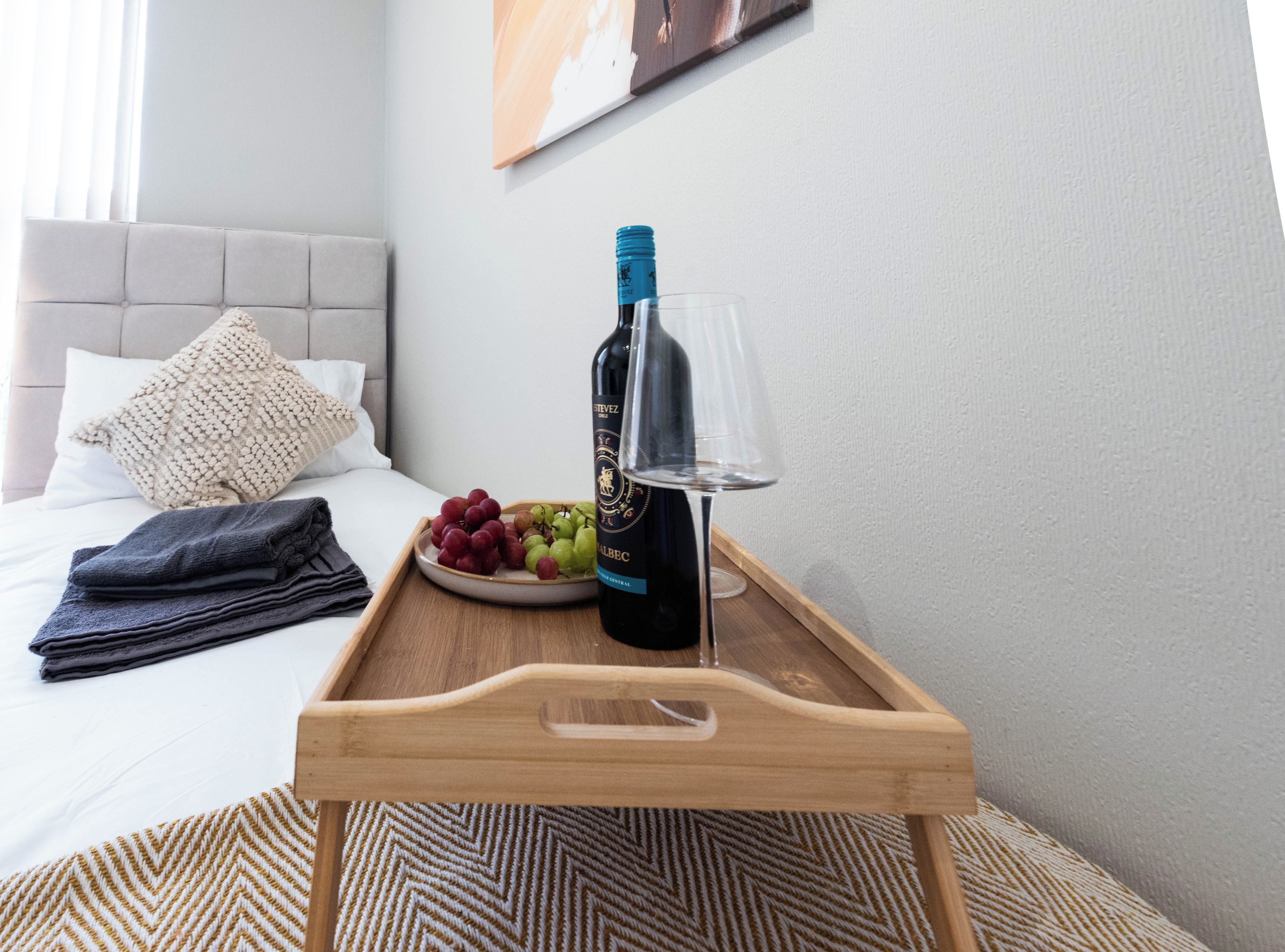Un lit avec une tête de lit, une table de chevet sur laquelle se trouve un plateau avec une bouteille de vin, un verre et un bol de raisins.