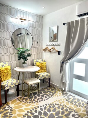 A room with a patterned wallpaper, a round mirror, a small round table, two chairs with yellow cushions, a potted plant, a wall-mounted air conditioner, a painting, and a window with curtains.