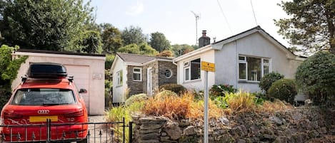 Une voiture rouge est garée devant une maison dont la façade est ornée d’un mur en pierre et qui est bordée d’un petit jardin.