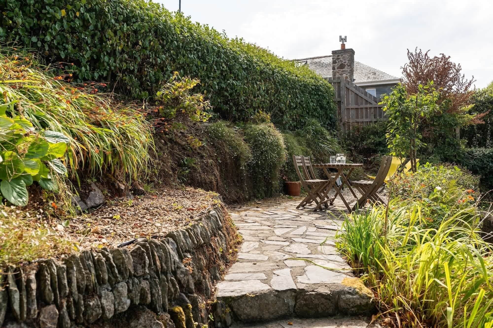 Un vialetto in pietra conduce a un tavolo e sedie di legno in un giardino con fitta vegetazione e un muro di pietra.