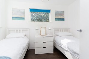 A bedroom with two single beds, a dresser with drawers, a mirror, bedside lamps, and two wall-mounted paintings. - Modern Home & Garden with Parking, Gorran Haven (Gorran Haven)