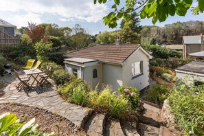 A small house with a sloped roof, surrounded by a garden with various plants and a patio area with outdoor furniture. - Modern Home & Garden with Parking, Gorran Haven (Gorran Haven)
