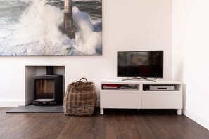 A modern living room with a fireplace, a television on a white cabinet, and a large painting of a lighthouse. - Modern Home & Garden with Parking, Gorran Haven (Gorran Haven)