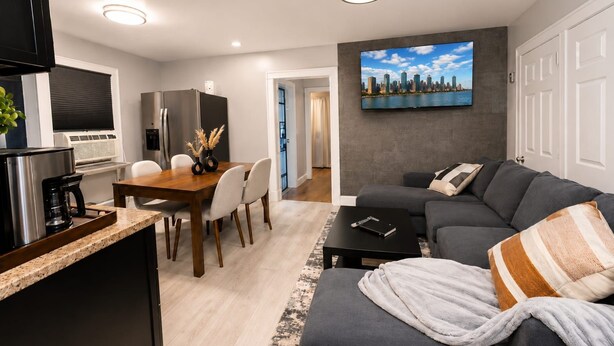 A modern living room with a grey sofa, a coffee table, a dining area with a wooden table and chairs, a kitchen with a refrigerator and stove, and a TV mounted on the wall. - Lovely 6-bedroom apartment with AC in vibrant Queens (Queens)