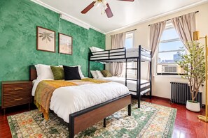 Ein Hotelzimmer mit Etagenbett, einem großen Bett mit gemustertem Teppich, einem Nachttisch, einem Fenster mit Vorhängen und einem Heizkörper.